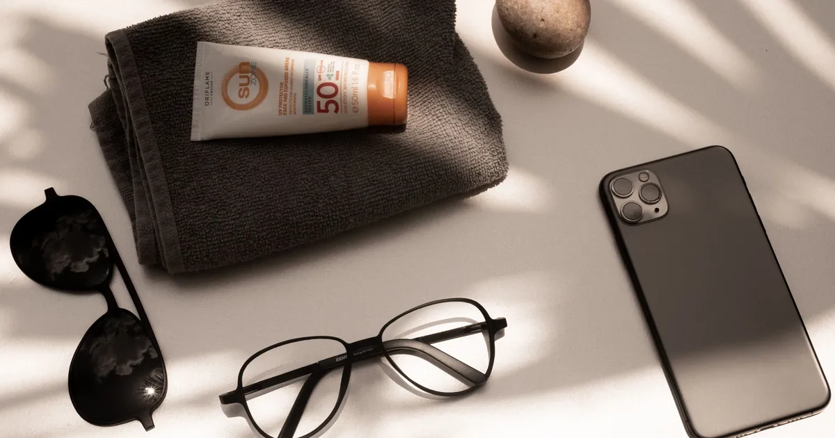 A tube of SPF sunscreen on a beach towel beside sunglasses and a phone